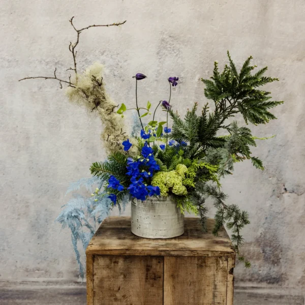 A sculptural winter floral arrangement with frosted evergreens, cobalt blue delphiniums, green hydrangeas, and natural branches in a white vessel. Perfect for Hanukkah.