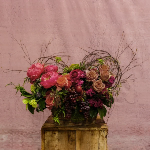 Aurora Symphony Basket featuring coral peonies, golden roses, magenta blooms, cascading grapes, and lush greenery in a woven basket.