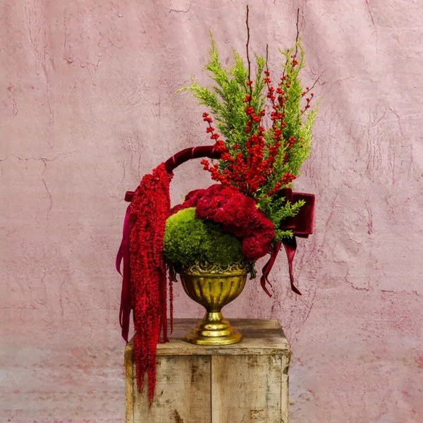 A festive Cerise Brilliance Basket featuring cerise-red celosia, green moss, ilex berries, cascading amaranthus, and a velvet ribbon in a gold pedestal bowl.