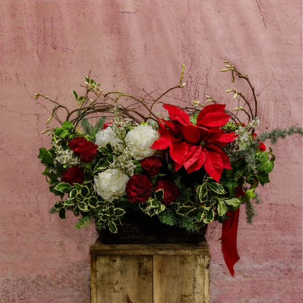 Christmas Charm Basket featuring red poinsettias, crimson carnations, white flowers, greenery, and willow branches in a rustic woven basket.