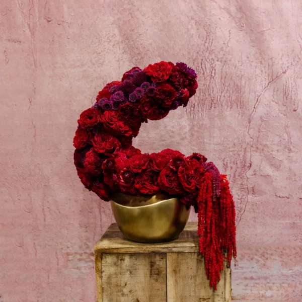 Crescent-shaped floral arrangement with red and burgundy roses, cascading amaranthus, and a gold vessel base.