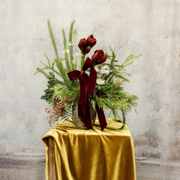 A holiday floral arrangement featuring burgundy amaryllis, white blooms, evergreen branches, and a velvet ribbon, perfect for winter or seasonal décor.