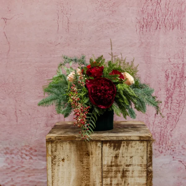 rimson floral centerpiece featuring burgundy peonies, blush roses, red ranunculus, pink pepper berries, and evergreen greenery in an emerald green vase, part of the Crimson Euphoria Collection.