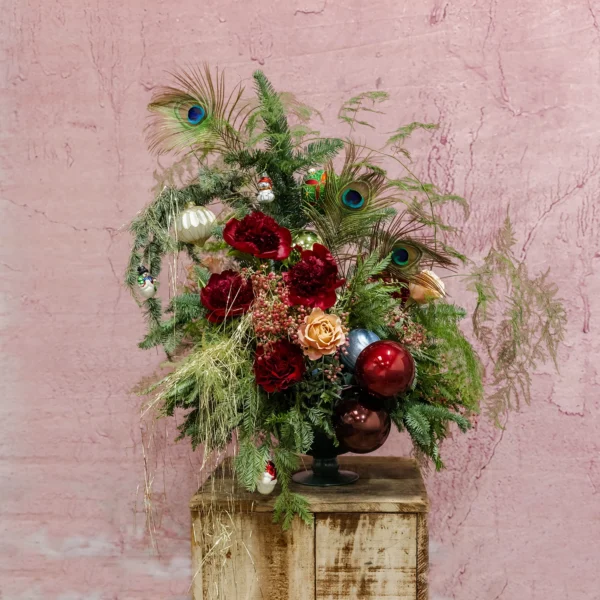 A festive arrangement featuring red blooms, blush accents, lush greenery, shimmering ornaments, and playful feathers, part of the Joyeux Radiance Collection.