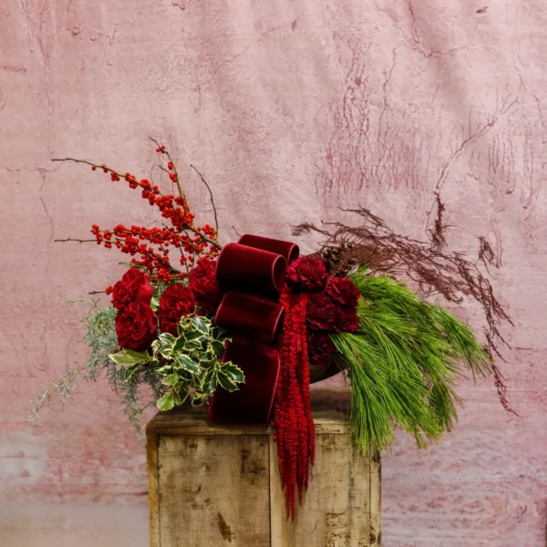 Festive floral centerpiece featuring red blooms, pine boughs, winter berries, and a velvet ribbon.