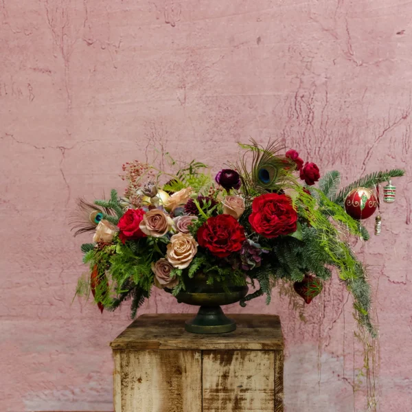 A centerpiece featuring vibrant red blooms, blush accents, greenery, ornaments, and peacock feathers, part of the Joyeux Radiance Collection.