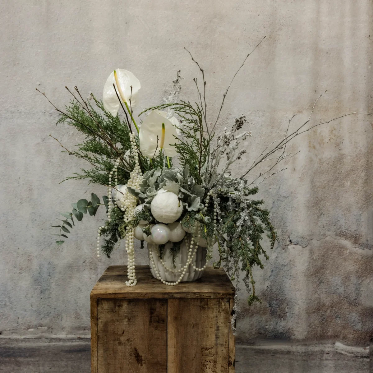 A winter floral arrangement with white anthuriums, roses, dusty miller, frosted greenery, eucalyptus, pearl garlands, and opalescent ornaments, ideal for holiday or seasonal décor.