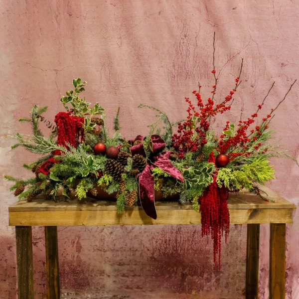 A festive centerpiece featuring crimson ilex berries, frosted holly, cascading amaranthus, pinecones, red ornaments, and a velvet ribbon, perfect for holiday decor.