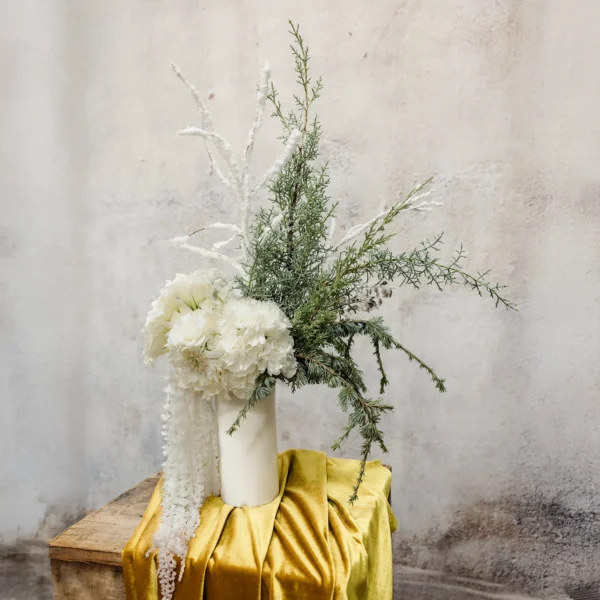 A winter bouquet with white flowers, frosted branches, evergreen accents, and cascading details in a minimalist white vase, perfect for holiday décor.