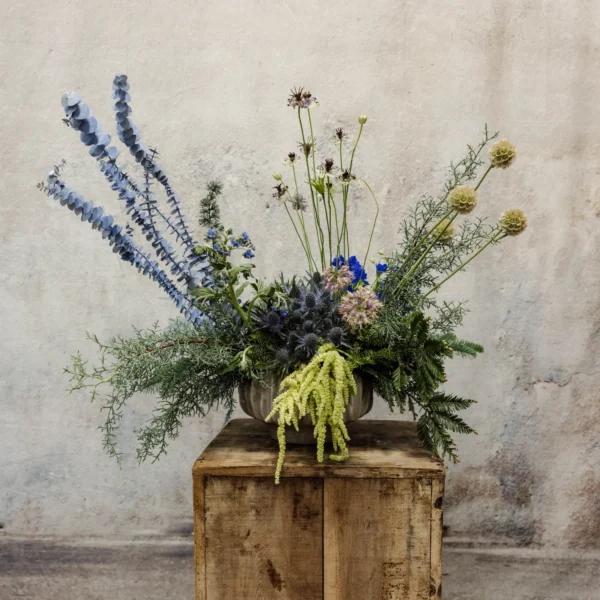 A dynamic floral arrangement featuring eucalyptus, cobalt blue flowers, thistles, seedpods, cascading amaranthus, and textured evergreens, perfect for Hanukkah or winter displays.