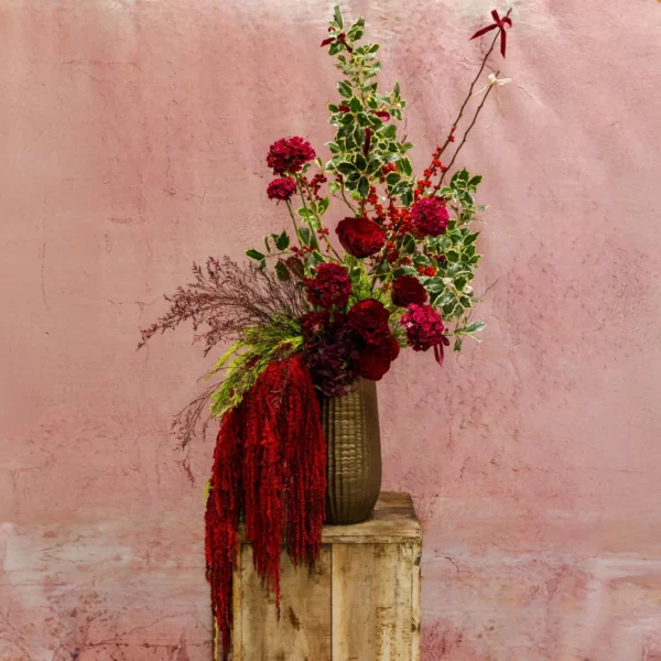 Tall floral arrangement with deep red blooms, cascading crimson accents, and lush greenery in a metallic vase, creating a dramatic seasonal display.