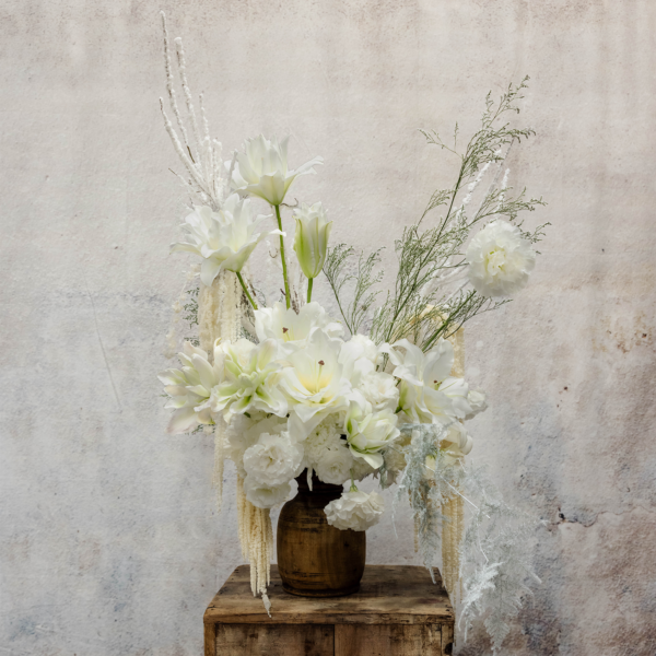 A tall winter floral arrangement featuring white casa blanca lilies, roses, ranunculus, frosted greenery, and cascading accents in a rustic vessel, perfect for holiday décor.