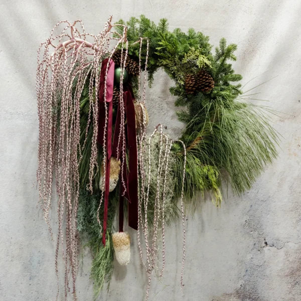 A holiday wreath featuring evergreen branches, pinecones, pink beaded accents, and a burgundy ribbon, perfect for rustic or seasonal décor.