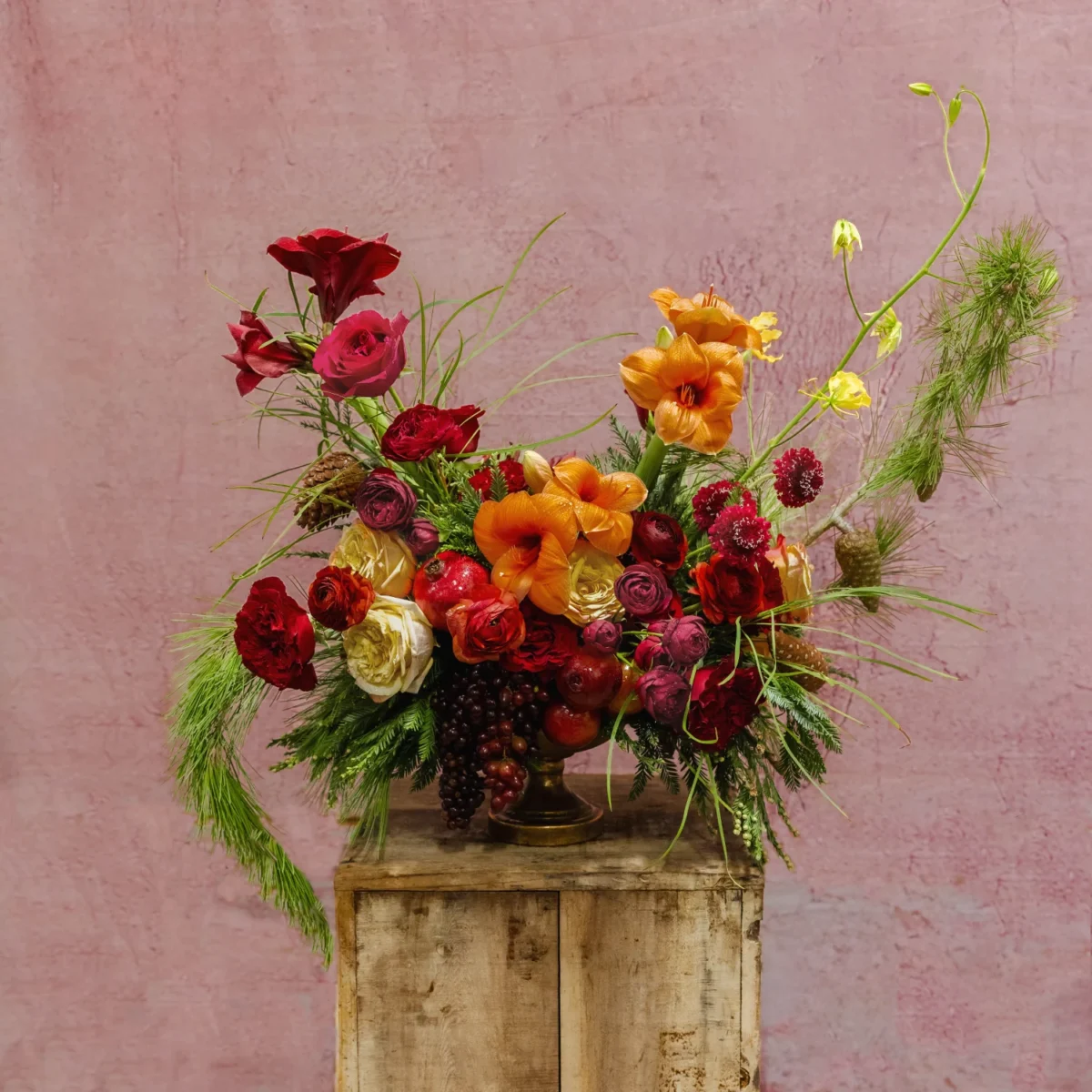 A festive arrangement featuring amber gladiolus, red ranunculus, blush roses, greenery, and seasonal fruit accents, part of the Celestial Yule Collection.