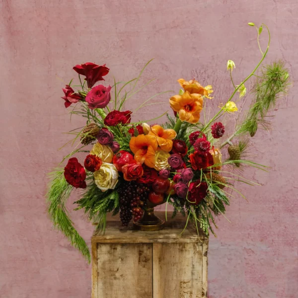 A festive arrangement featuring amber gladiolus, red ranunculus, blush roses, greenery, and seasonal fruit accents, part of the Celestial Yule Collection.