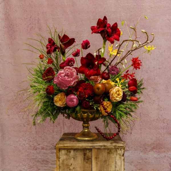 A centerpiece featuring deep red amaryllis, blush roses, pink peonies, golden accents, and lush greenery, part of the Celestial Yule Collection.