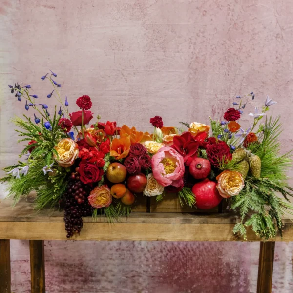 A centerpiece featuring red and blush blooms, orange accents, pomegranates, grapes, and lush greenery, part of the Celestial Yule Collection.