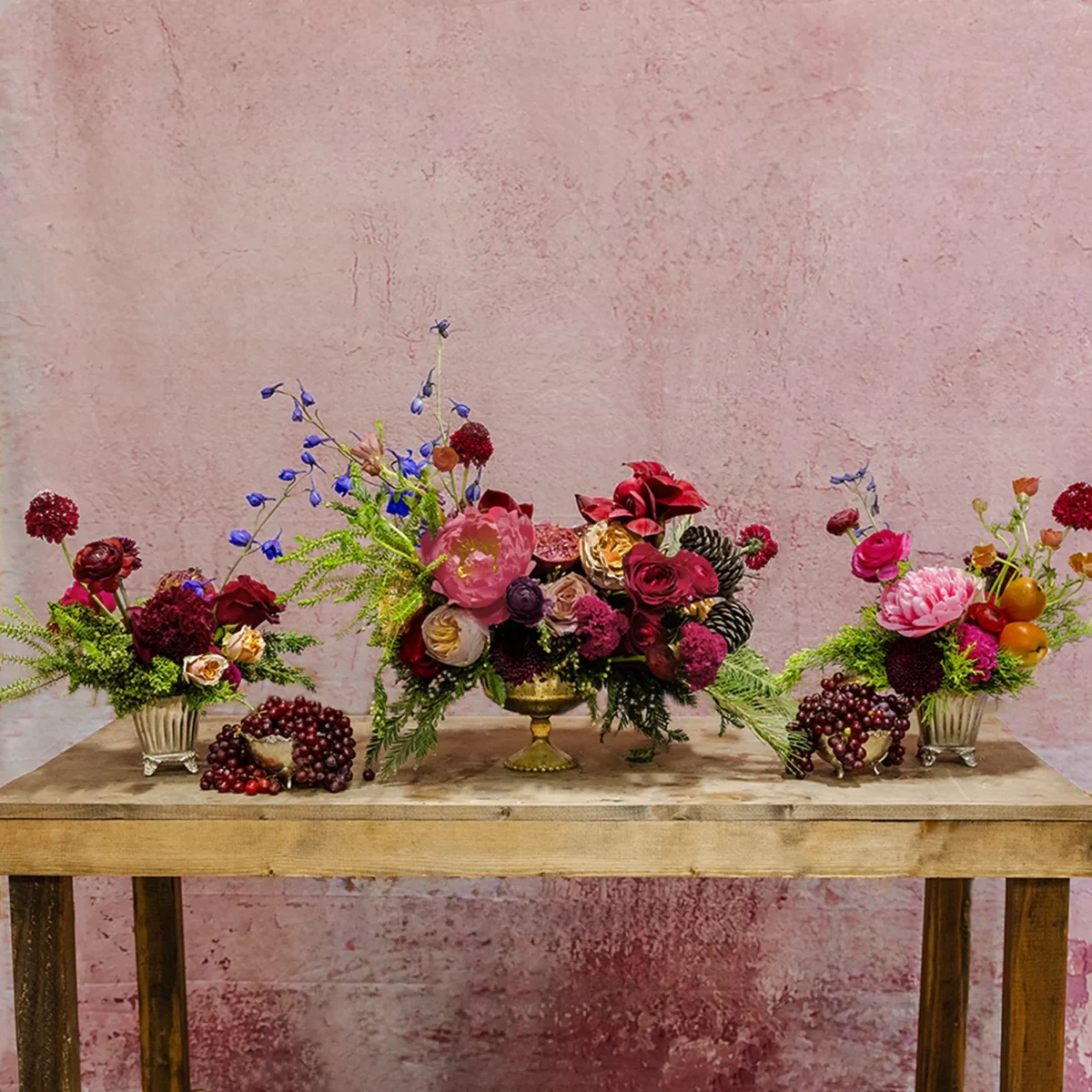 A trio of holiday centerpieces featuring red, pink, and blush blooms, lush greenery, grapes, pomegranates, and cobalt blue accents, part of the Celestial Yule Collection.