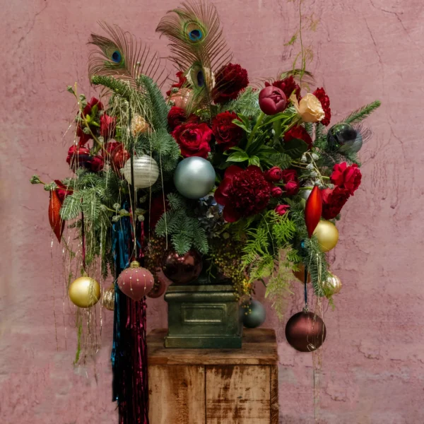 A luxurious centerpiece with deep red blooms, lush greenery, shimmering ornaments, cascading ribbons, and peacock feathers, part of the Joyeux Radiance Collection.