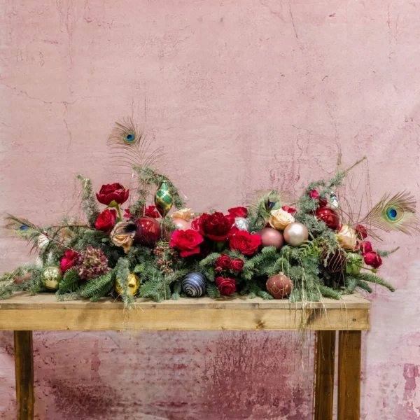 A festive holiday centerpiece with evergreen boughs, red roses, blush accents, shimmering ornaments, and peacock feathers, part of the Joyeux Radiance Collection.