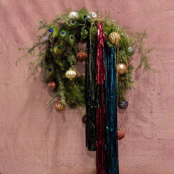 A festive holiday wreath with lush greenery, vibrant feathers, sparkling ornaments, and cascading jewel-toned ribbons, part of the Joyeux Radiance Collection.