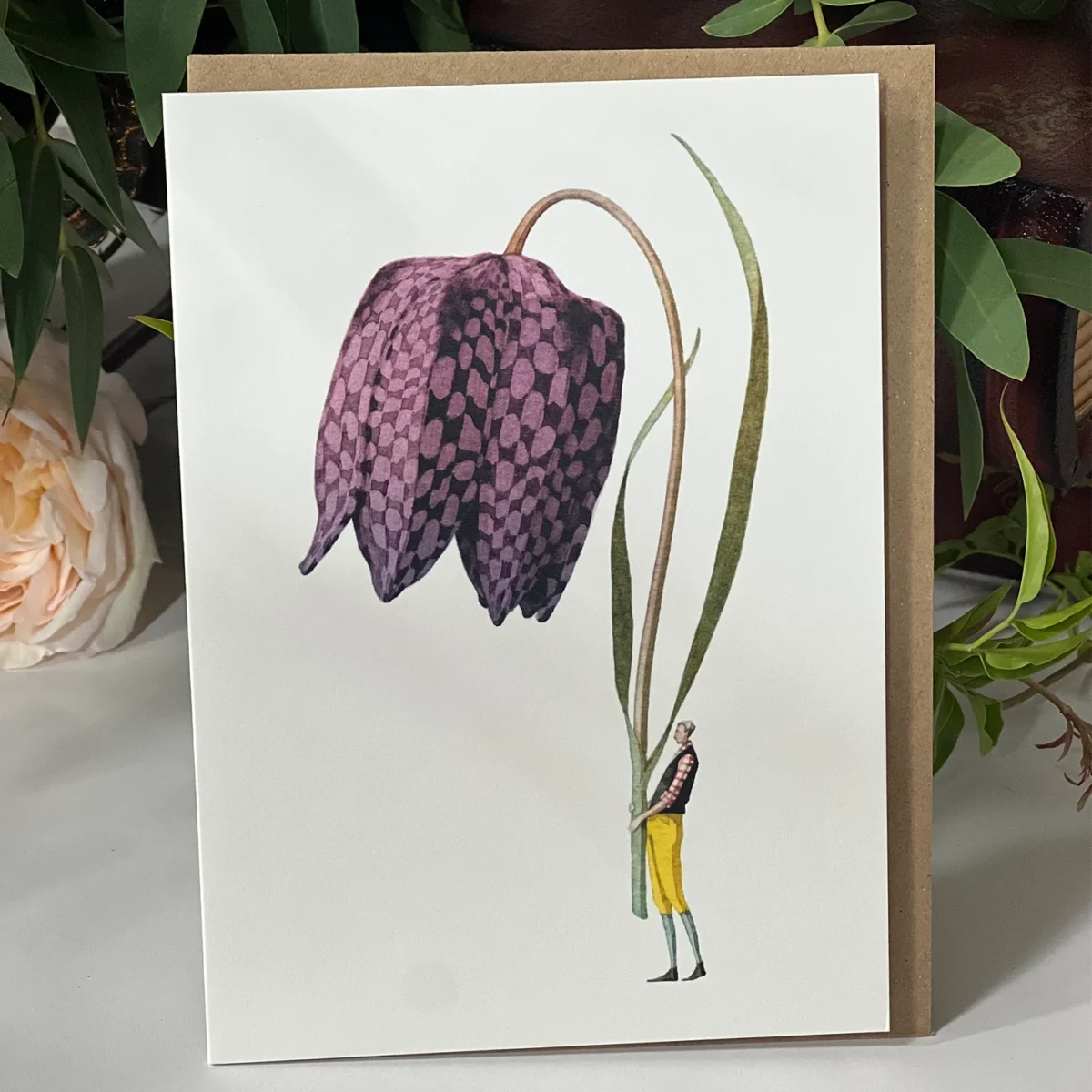 Illustrated greeting card with a purple checkered flower held by a small figure wearing yellow pants, surrounded by greenery.