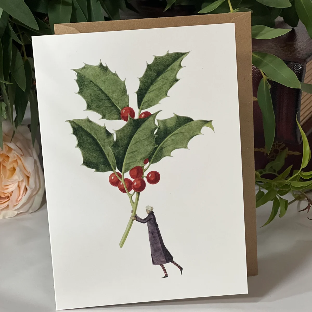 Greeting card depicting a festive holly branch with green leaves and red berries held by a small illustrated figure in a dark coat