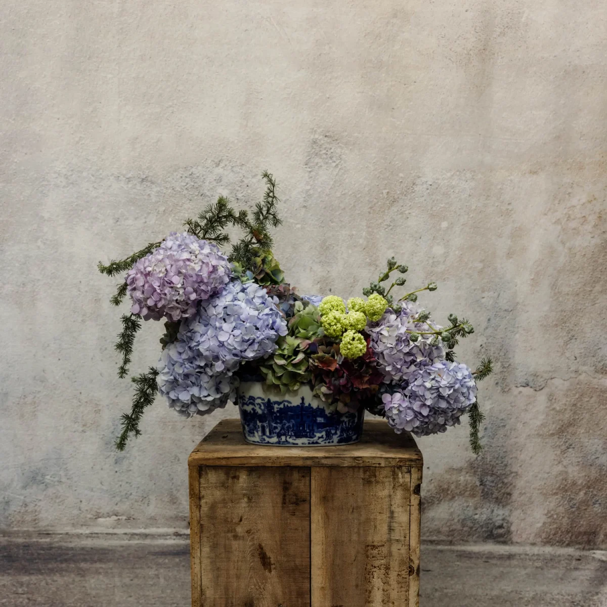 A sophisticated arrangement of blue and green hydrangeas with viburnum and evergreen sprigs, presented in a blue-and-white ceramic vessel, part of the Luminous Heritage Collection.