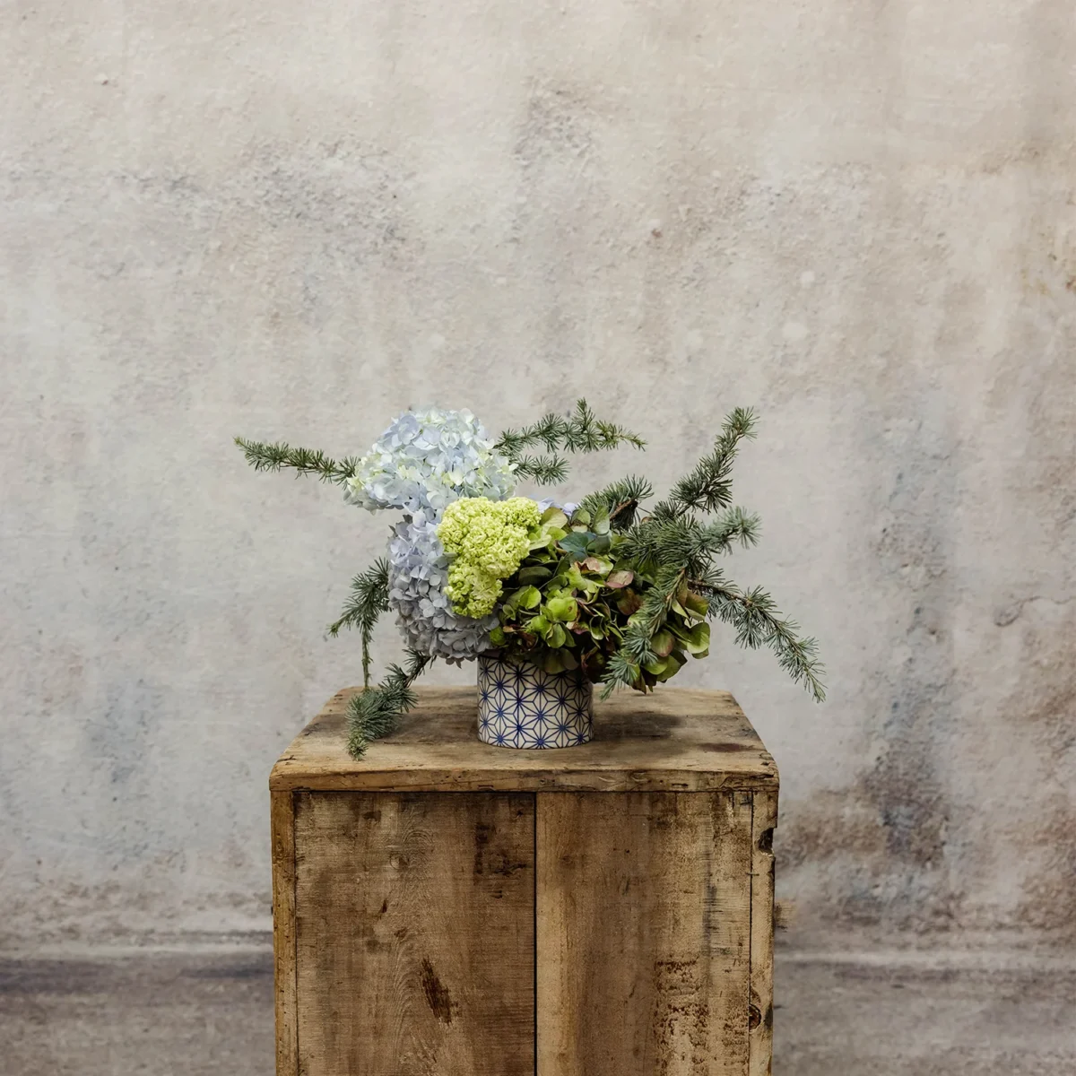 A beautiful arrangement of blue and green hydrangeas with viburnum and evergreen branches, styled in a patterned ceramic vase, part of the Luminous Heritage Collection.