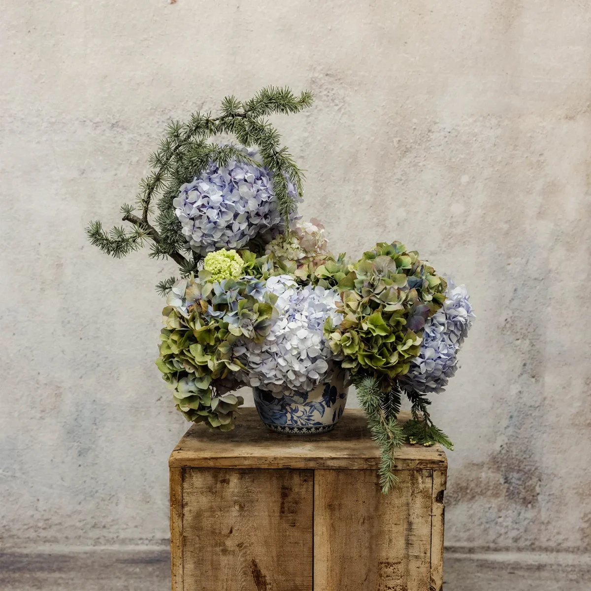 A refined winter floral arrangement featuring blue and green hydrangeas, viburnum blooms, and evergreen sprigs in a blue-and-white ceramic vase, ideal for seasonal or home décor.